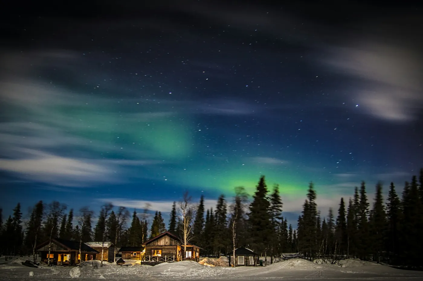 Wooden cabins with lit windows at the edge of a spruce forest, green aurora and drifting clouds in the starry sky above