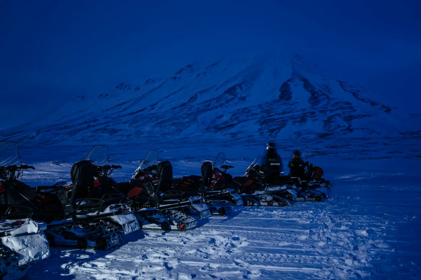 A row of parked snowmobiles on packed snow below a snow-covered mountain, everything in deep blue twilight