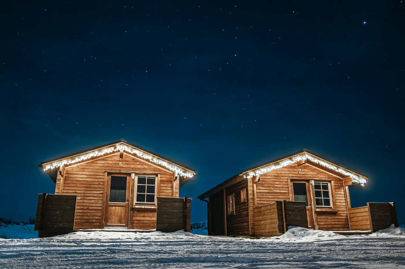 Two small wooden cabins with string lights along the roofs, standing in snow under a dark blue starry sky