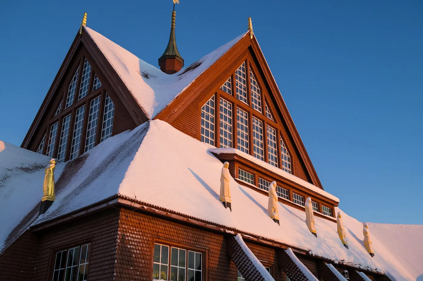 Steep snow-covered roof and gables of a dark red wooden church with tall windows and small gilded figures along the eaves, under a clear blue sky