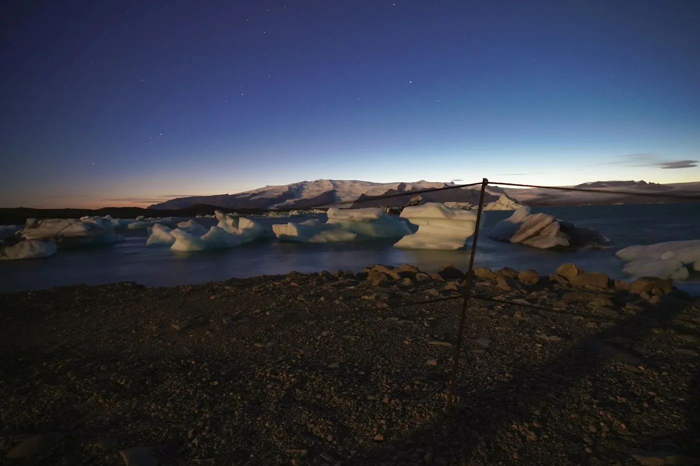 Blocks of ice floating on dark water below snowy hills, with a faint orange glow on the horizon and stars above