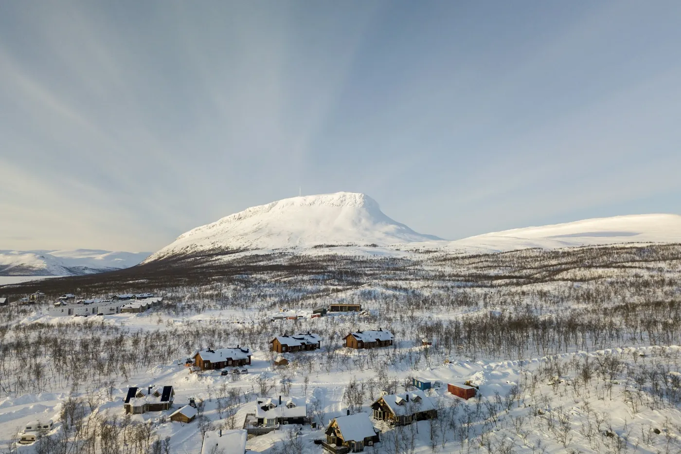 A snow-covered rounded fell above scattered wooden cabins and low birch woodland under a pale winter sky