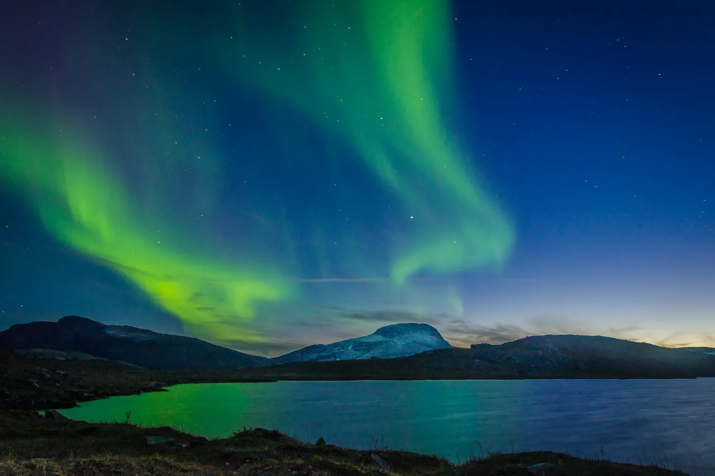 Green northern lights over a lake and a flat-topped mountain, a band of blue twilight on the horizon