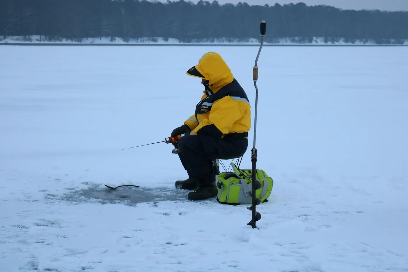 A person in a yellow and black winter suit sits on a stool on a frozen lake, fishing through a hole in the ice, with an ice drill standing beside them