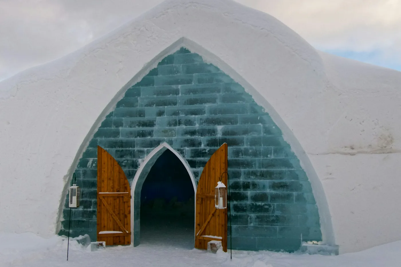 Pointed entrance arch built from blocks of clear ice in a snow wall, with two open wooden doors and lanterns on either side