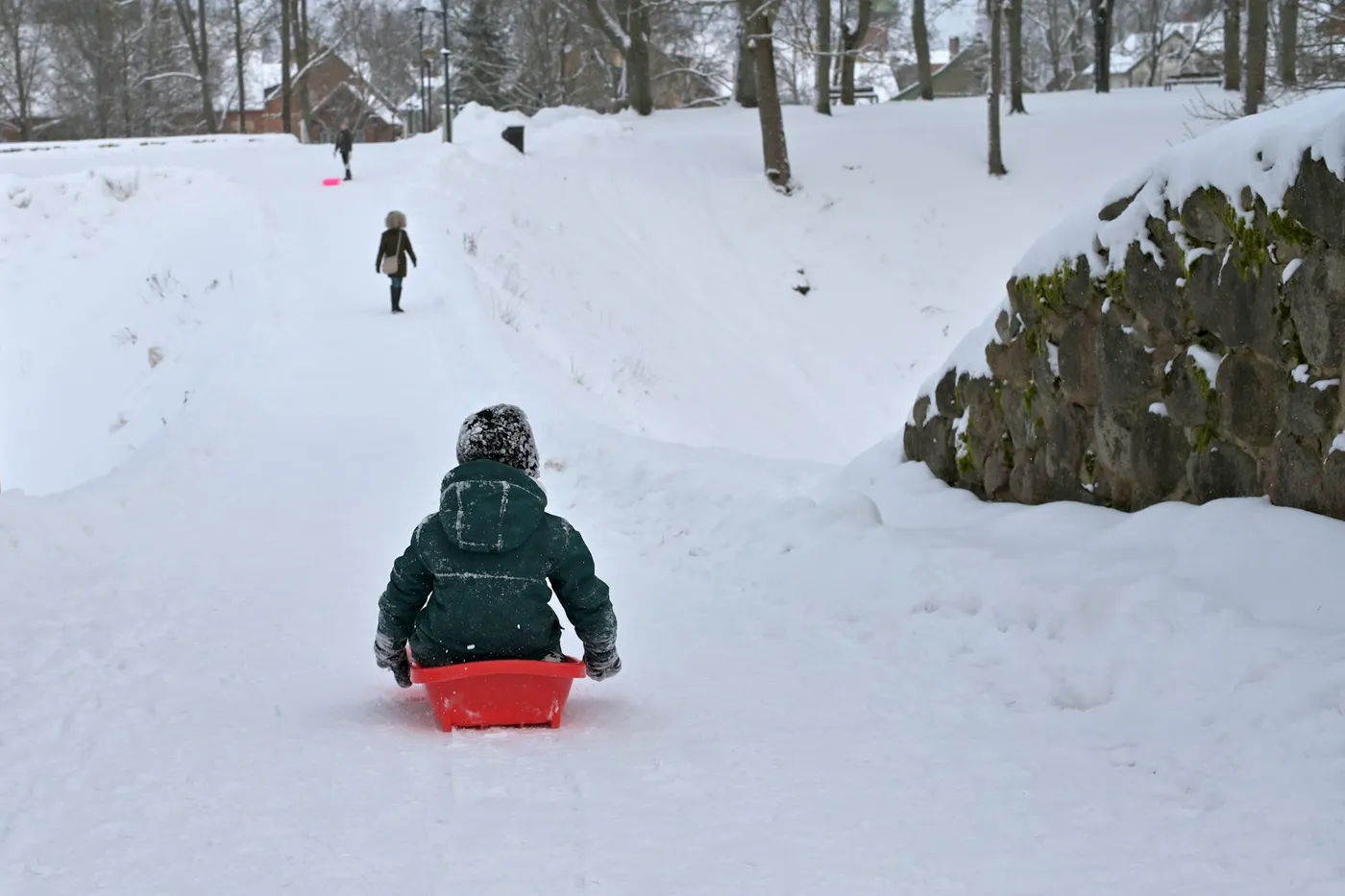 A small child in a green winter jacket and knitted hat sliding down a snowy path on a red plastic sled, two people walking further up