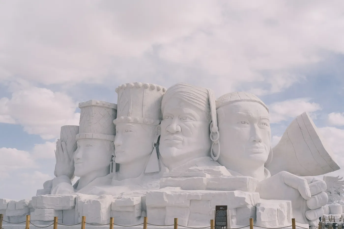 A large white snow sculpture of four faces wearing different headwear, behind a low rope fence under a cloudy sky