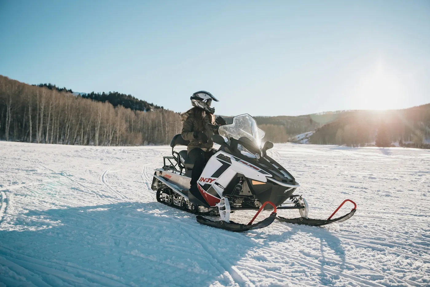 A rider in a helmet sitting on a parked snowmobile on a snowy field, low sun behind a wooded hill