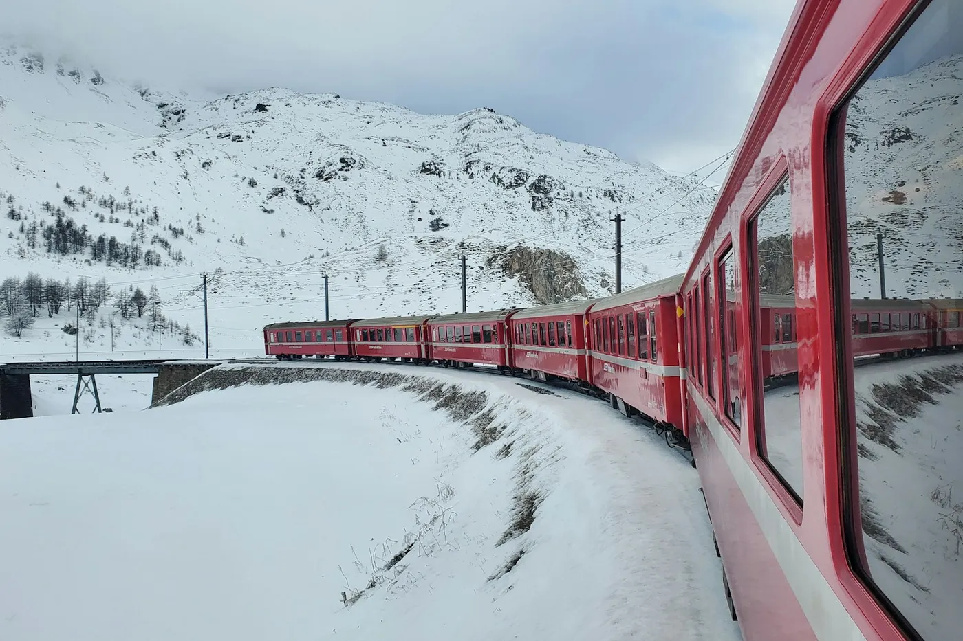 A red passenger train curving through a snowy valley below white mountains, seen from one of its windows