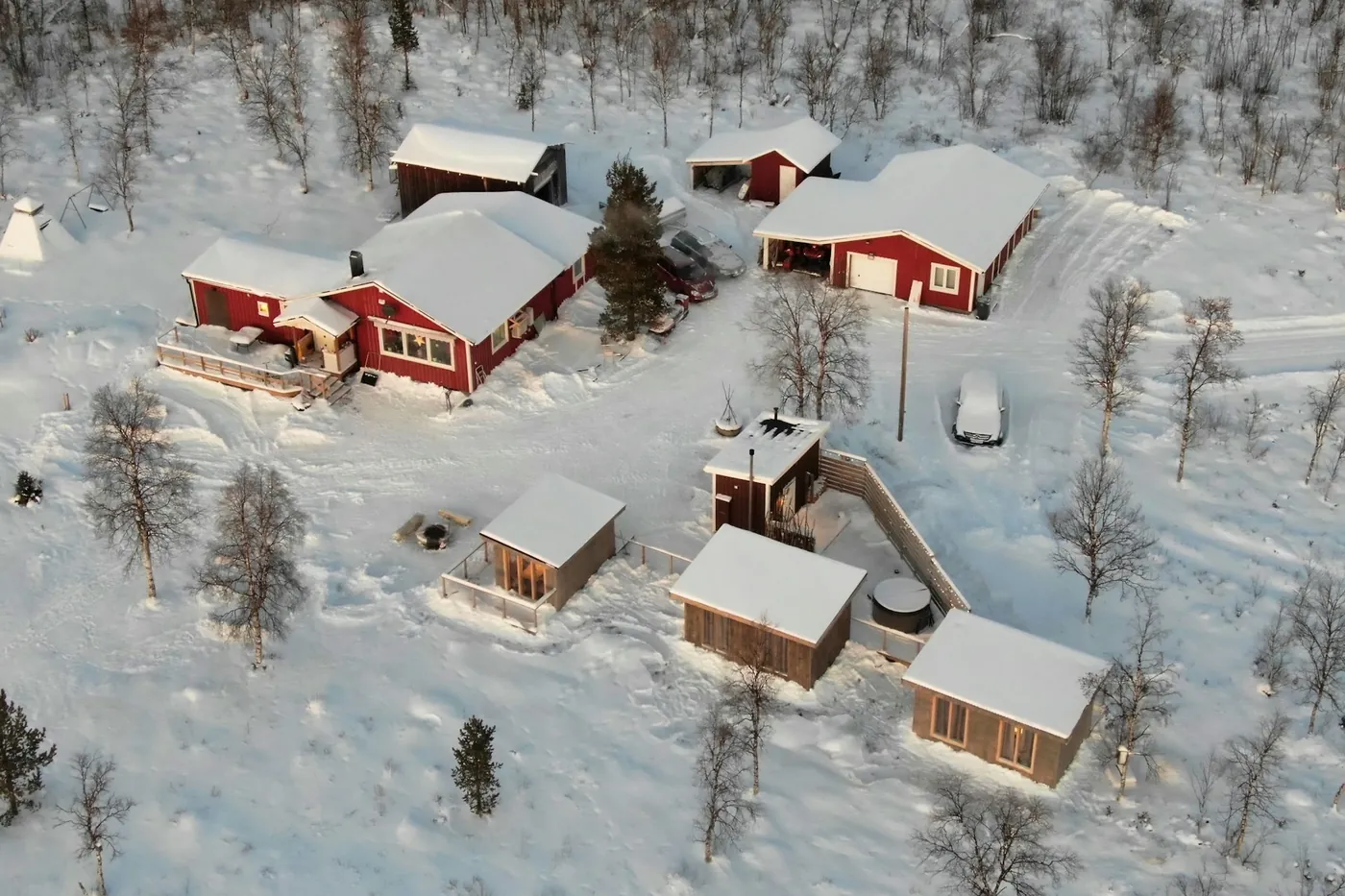 Aerial view of red wooden houses, cabins and a small tent-shaped hut in deep snow, birch trees around them